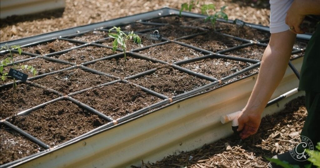 A person adjusts a knob on a raised garden bed with a square grid marking planting sections and young plants, helping their garden survive summer.