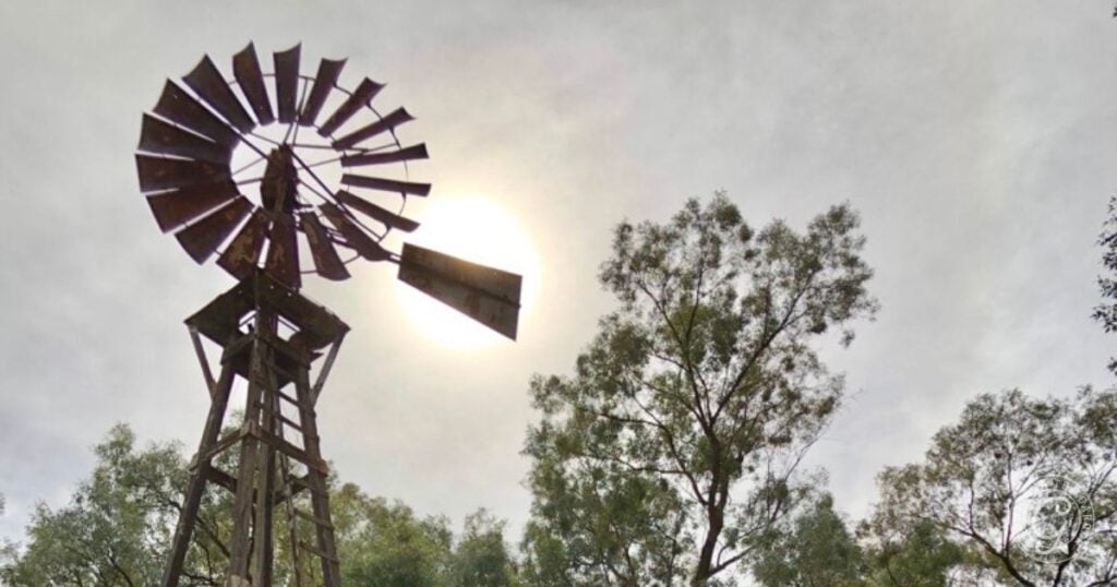 A metal windmill stands tall among trees in a garden, enduring the sun shining through a cloudy sky behind it—ready to survive summers heat.