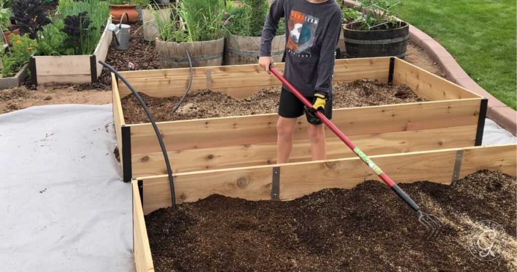 A child rakes soil inside a wooden raised garden bed in a backyard garden, preparing the area as part of learning how to kill Bermuda Grass for healthier plants.